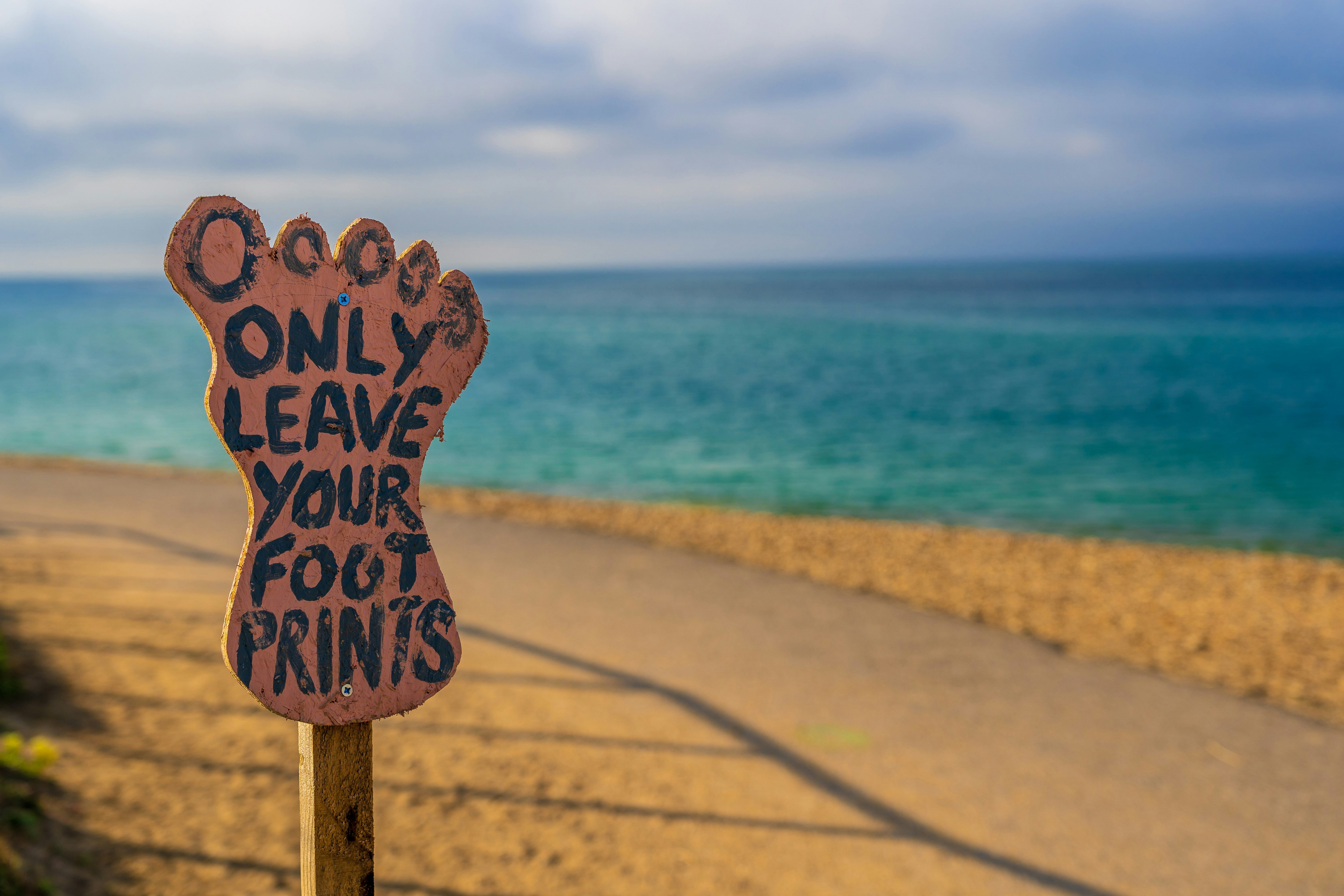 A primitive wooden sign in the shape of a foot on a beach with the words 'Only leave your foot prints' painted on it.